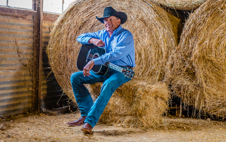 George Strait sitting on a hay bail holding a guitar.